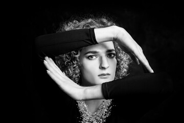 Black and white portrait of a sad androgyne guy model with blue eyes and blond hair in the image of a girl with beads around neck, holds hands with a frame in front of his face.