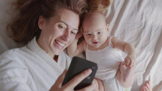 Close up of mother and her newborn baby making a selfie or video call to father or relatives in a bed. Concept of technology, new generation, family, connection, parenthood.