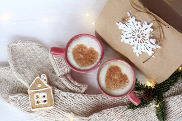 Obraz na płótnie Canvas two mugs with a pair of hearts on a frothy surface cappuccino and a gingerbread house with a gift, mittens and a scarf on the table top view. warm moments in the day of lovers
