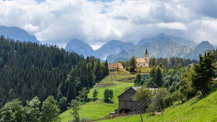 Fototapeta na wymiar Church Cerkev Sveti Duh in Solčava with mountain range Kamnik–Savinja Alps in Slovenia on a sunny summer day.