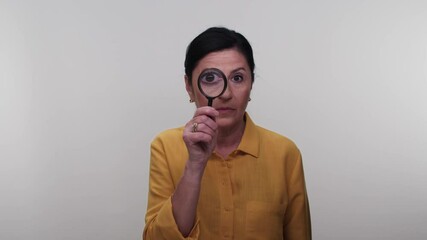 Elderly woman with magnifying glass looking at camera.The old lady is holding a magnifying glass on a white background and looking through the eye. Smart People Concept, Researchers. Close up. - Powered by Adobe