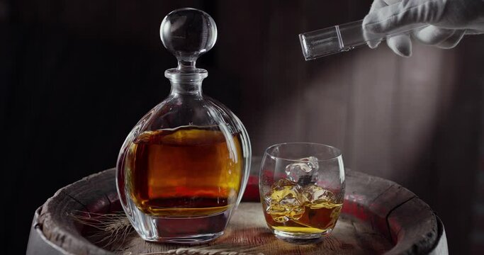 An ice cube slowly falls into a glass of whiskey on top of a vintage whiskey barrel. A decanter with a drink stands nearby, a dark brown background. Blackmagic Ursa Pro G2