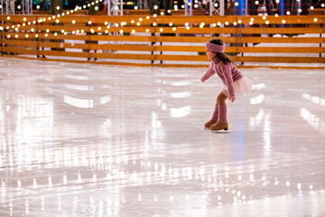 Little girl figure skater in pink sweater is skating on winter evening on an outdoor ice rink lit...