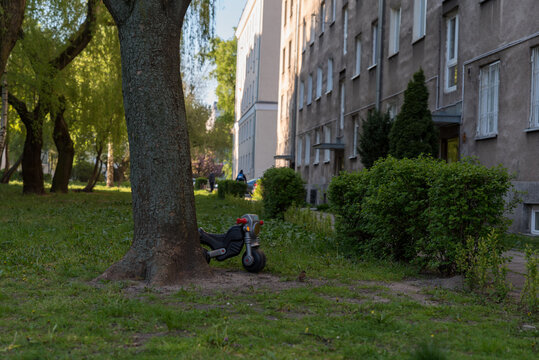 Spring In The City, A Children's Toy Motorcycle Left On The Green Lawn Behind A Tree. Toy In Front Of The Block.