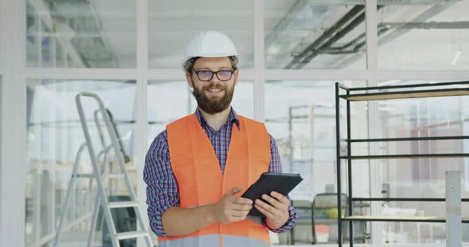 Handsome positive satisfied skilled bearded builder in hardhat,gogglesand vest using tablet pc in new built office and looking at camera with sincerelt smile