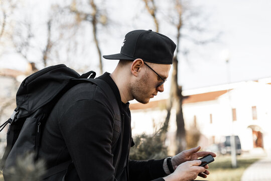 American good-looking young man hipster in sunglasses in black clothes in fashion baseball cap with backpack with mobile phone sits on street. Handsome urban guy fashion model is looking in smartphone