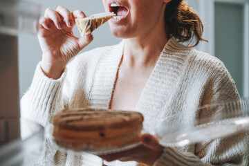 Attractive friendly brunette woman middle age in cozy cardigan eating cake from refrigerator at the...