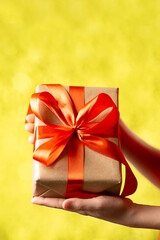 Hands Holding Craft Gift Box With Red Ribbon on Abstract Bokeh Background. Vertical shot.