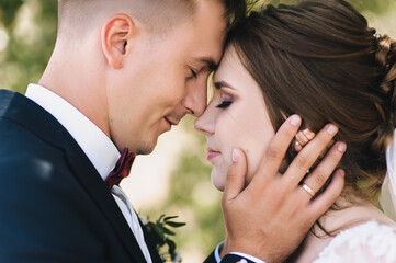 Wedding portrait of the newlyweds, close-up. Stylish groom in a blue suit and a beautiful curly-haired bride embrace, standing with their foreheads close.