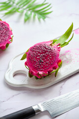 Pink dragon fruit, pitaya or pitahaya cut in cubes on white plate. Trendy superfood ingredient. White table with cutting board, knife and exotic palm leaves.