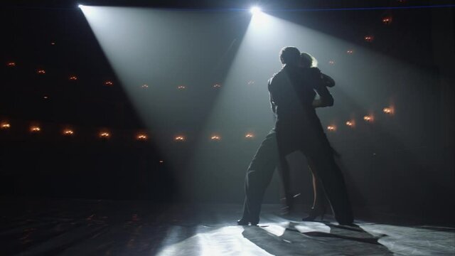 Attractive couple of ballroom dancers dancing passionate latin american dance cha cha cha. Silhouettes of two dancers on a smoky Stage with spot light. Anamorfic lens