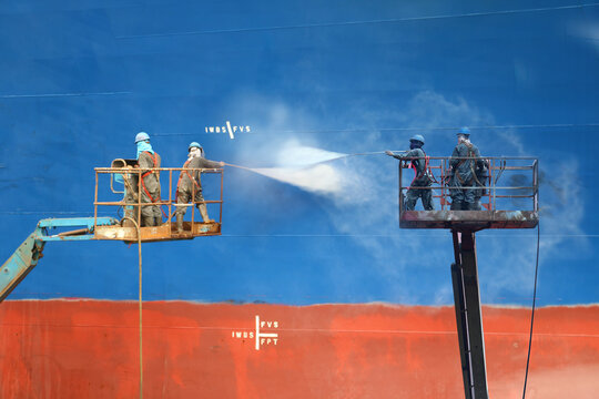 Shipyard worker power washing, cleaning water jet high pressure a ship in floating dry dock on sherry picker car on high wearing safety harness both.