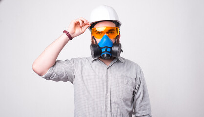 Obraz premium Young bearded engineer wearing a helmet, protective glasses and a respirator is looking at the camera near a white wall .