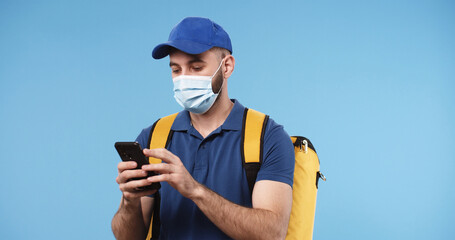 Portrait of delivery man in medical mask with yellow thermal backpack using smartphone isolated over blue background. Online shopping and delivery on quarantine.