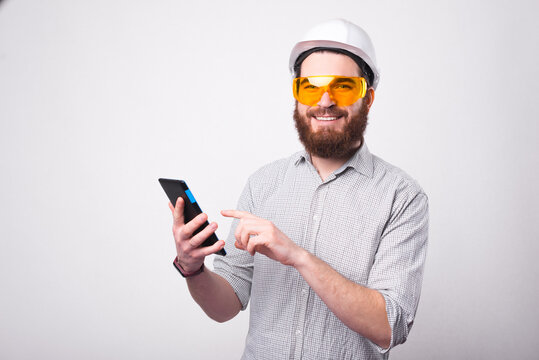 Cheerful bearded architect is holding a tablet and smiling at the camera is wearing a hemlet and a pair of protective glasses .