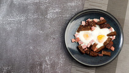 Fried eggs with dark rye bread and sausage on a round plate on a dark background. Top view, flat lay