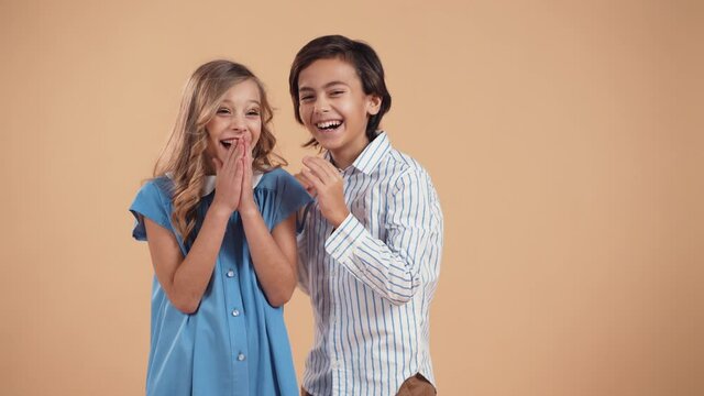 A cute boy in a striped shirt whispers a joke or gossip to a cute girl in the ear with curly hair in a blue dress and they laugh together isolated on a peach tender background. Valentine's Day.