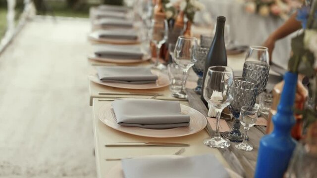 Table setting, textile napkins are on the plates, there are beautiful glasses and decorative bottles with flowers on the table, against the background of female hands putting napkins on the dishes