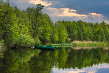 A river with trees around which are reflected in the water, a boat near the shore, and the sky with the setting sun that shines through the clouds.