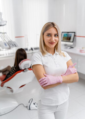 Blonde dentist doctor stands on patient in dentist chair background. Doctor in white scrubs at working place.