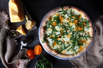 Pizza with several varieties of cheese and arugula. Traditional Italian pizza. Metal plate for serving pizza. A piece of cheese on a dark background. The view from the top.