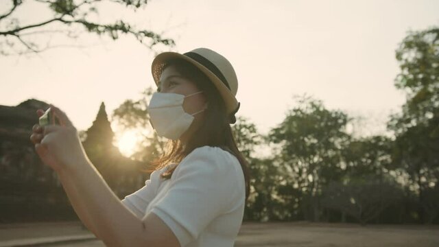 social distancing safety travel new normal lifestyle asian female woman wearing protective face mask walking and take photo with smartphone tour in public garden park sunset moment