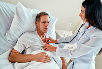 Obraz premium A beautiful young nurse with a stethoscope examines a mature patient who is lying on a bed in a modern ward.