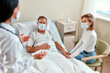Fototapeta na wymiar A beautiful woman doctor in a protective medical mask communicates with the family of a mature patient who is lying in a modern hospital ward during the coronavirus or covid-19 epidemic.