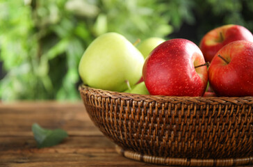 Different ripe apples in wicker bowl on wooden table against blurred background, closeup