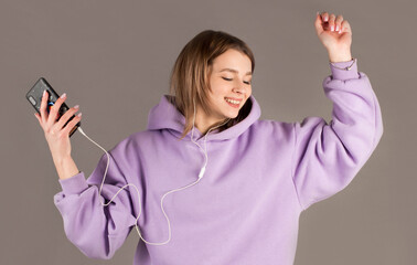 Happy carefree young woman dancing and listening to music from smartphone over grey background