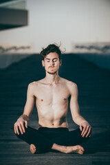 Young red-haired man practicing the lotus yoga posture, Padmasana, outdoors on the wooden floor