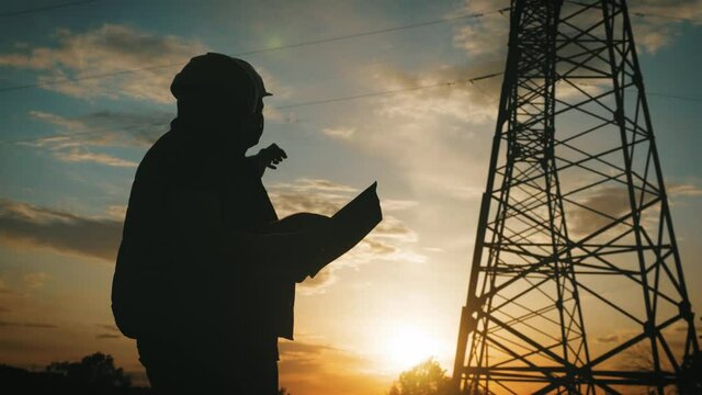 Silhouette of engineers looks at the construction of high-voltage power. Teams engineer looking discussing plan. Two engineer standing on field with electricity towers at sunset.