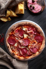 Pizza with ham, salami and hunting sausages.Traditional Italian pizza. Metal plate for serving pizza. A piece of parmesan and capers on a dark background. Vertical photo. The view from the top.