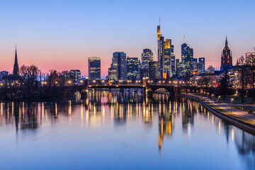 Fototapeta na wymiar Frankfurt skyline in the evening. Sunset at blue hour with illuminated skyscrapers from the financial and business district. Reflections on the river Main with park on the bank