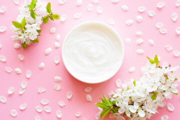 Fototapeta na wymiar Opened white cream jar on pastel pink table background. Beautiful cherry blossoms and petals. Care about clean and soft face, hands, legs and body skin. Fresh flowers. Top down view. Closeup.