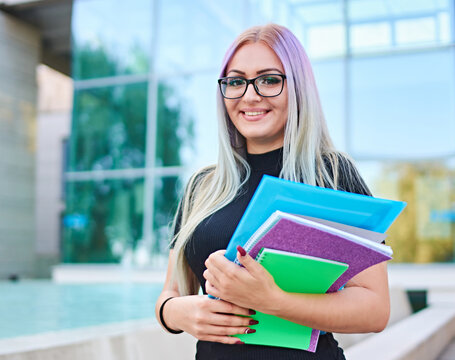 Young happy student girl standing in front of a campus building and looking into camera - Smart millennial woman with glasses holding booklets - Freshman with colorful hair smiling after an exam