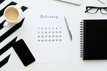 Calendar of February on white desktop flat lay with a cup of coffee and a notebook, work space