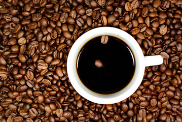 A cup of black coffee with two coffee beans floating in it stands against the background of many brown coffee beans