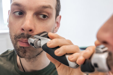 Close up portrait of man shaving beard or stubble with an electric razor. Reflection in mirror of...