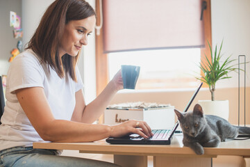 Young woman in white t-shirt sitting with a cat on her lap at the wooden table at home with laptop and notebook, working