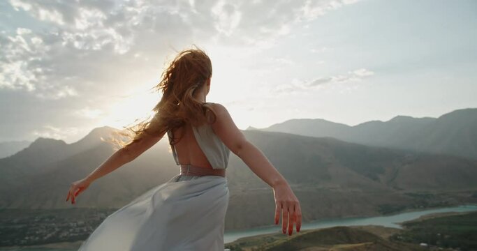 Woman in white dress standing on top of a mountain with raised hands while wind is blowing her dress and red hair - freedom, nature concept 4k footage
