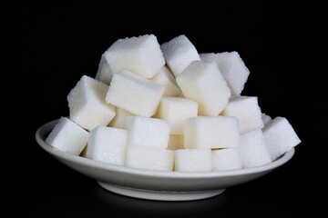 Sugar is white in a plate on a black background. It is used in food products and added to beverages.