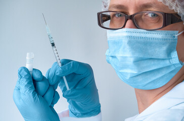 Coronavirus vaccine concept. Medic, doctor or nurse with covid-19 vaccine vial and syringe. Mature European female tech in face mask, glasses, hat and blue gloves looks at camera.