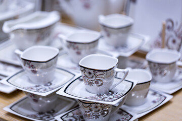 A set of plates and cups in white tone stands on a light wooden table. A stack of ceramic plates, glasses, mugs. Simple design, close-up top view. Place for your text. empty mugs of tea and saucers.