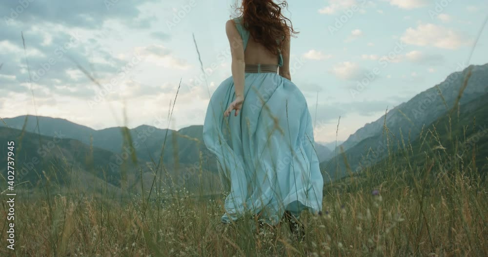 Wall mural Woman in white dress running on a mountain with raised hands while wind is blowing her dress and red hair - freedom, nature concept 4k footage