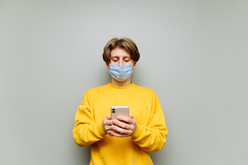 Serious guy in a medical mask uses a smartphone on a background of a gray wall, looking intently at the screen. Guy in a protective mask uses the Internet on a smartphone