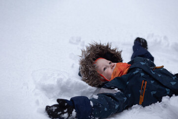 Fototapeta na wymiar Cute smiling toddler girl wearing winter clothes is lying on the snow and creating snow angel. Copy space