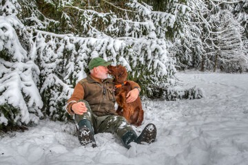 Fototapeta na wymiar Close friends. In the snowy winter forest an elderly hunter sits in the snow with his young Irish Setter hunting dog and is tenderly bitten in the nose..