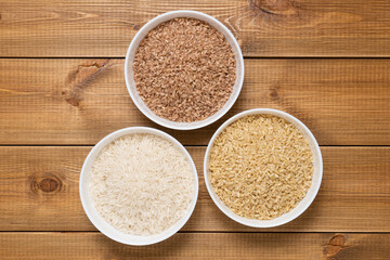 Raw uncooked rice in white ceramic bowls on wooden table, top view. Brown, red and white long basmati rice varieties. Gluten free products.