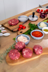 Burger press on wooden cutting board, vegetables and spices with kitchen bowls.